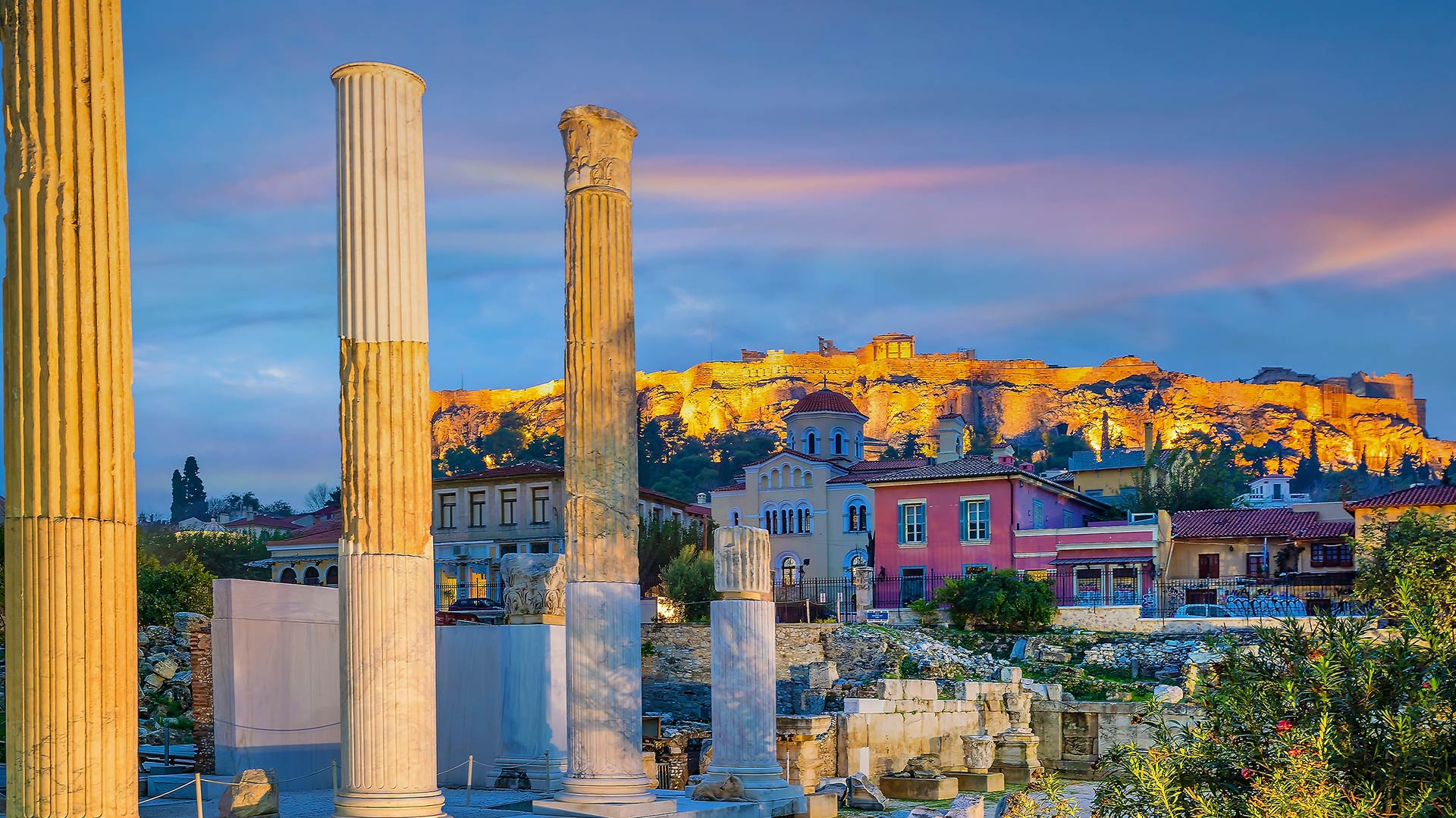 Ancient columns and ruins in Athens at sunset with the illuminated Acropolis in the background, showcasing the city's rich historical and cultural heritage.