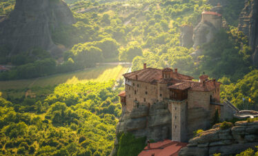 Monastery perched atop dramatic rock formations in Meteora, surrounded by lush green valleys and golden sunlight.