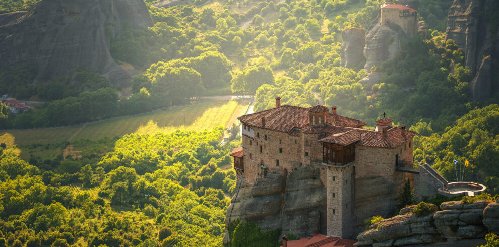 Monastery perched atop dramatic rock formations in Meteora, surrounded by lush green valleys and golden sunlight.