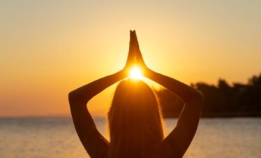 Woman practicing yoga with hands raised toward the setting sun on a serene beach in Lesvos, Greece.