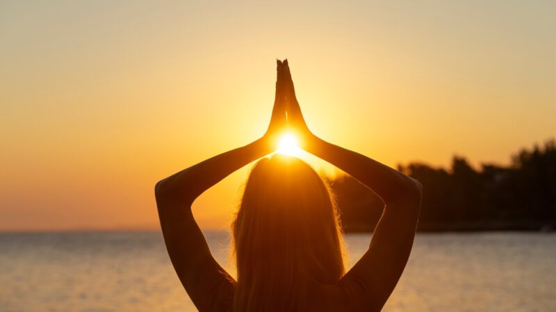 Woman practicing yoga with hands raised toward the setting sun on a serene beach in Lesvos, Greece.