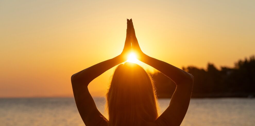 Woman practicing yoga with hands raised toward the setting sun on a serene beach in Lesvos, Greece.