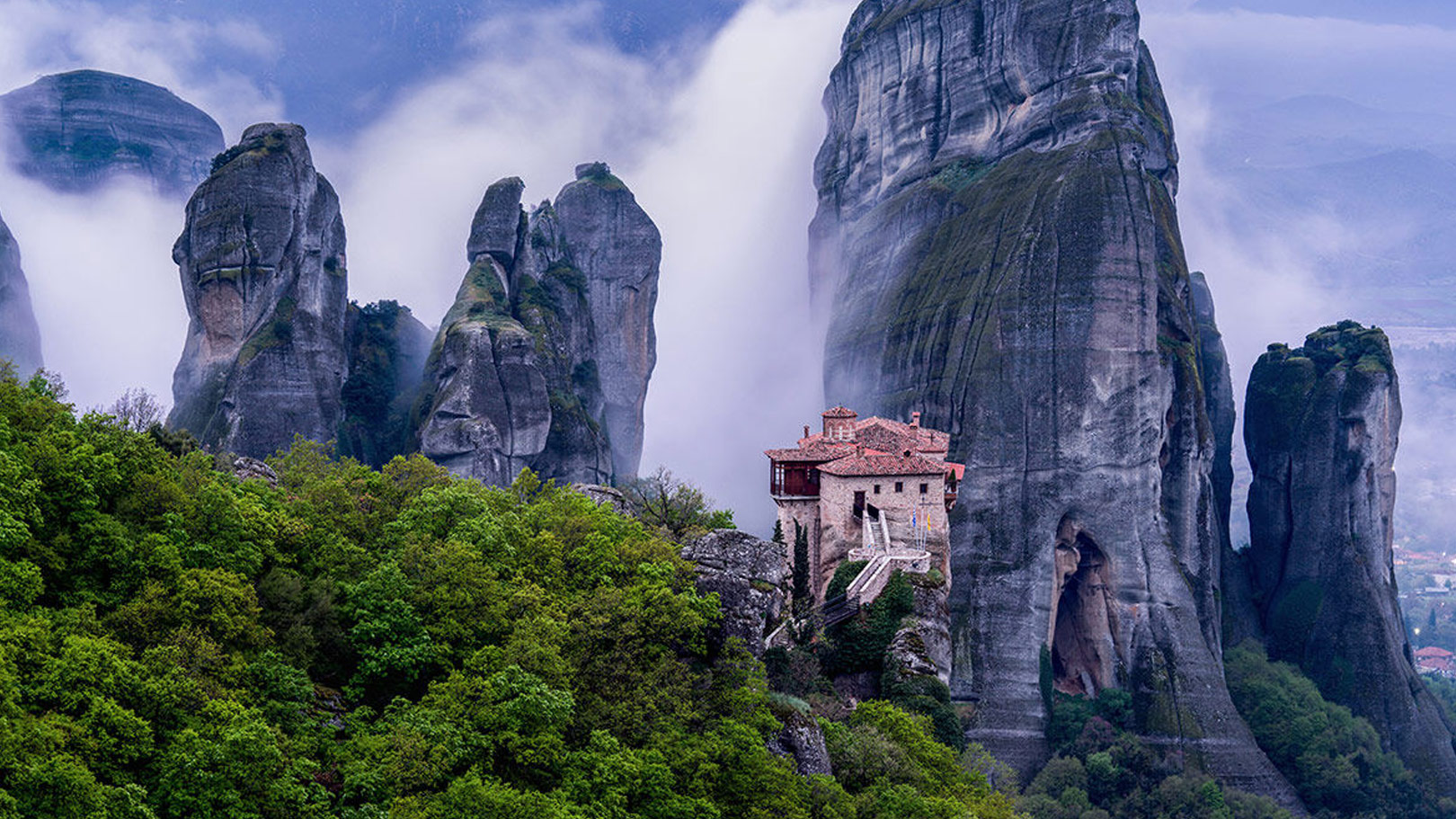 Monastery perched on towering rock formations surrounded by mist and lush greenery in Meteora, Greece.