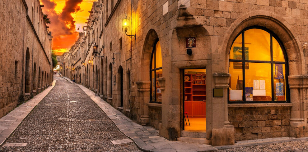 Sunset view of the medieval Street of the Knights in Rhodes Old Town with historic stone buildings and warm lights.