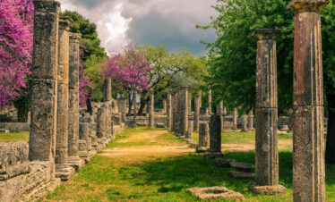Ancient ruins of Olympia surrounded by blooming trees and greenery under a dramatic cloudy sky in Greece.