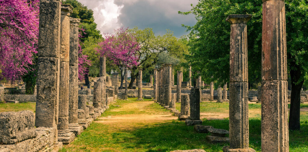 Ancient ruins of Olympia surrounded by blooming trees and greenery under a dramatic cloudy sky in Greece.