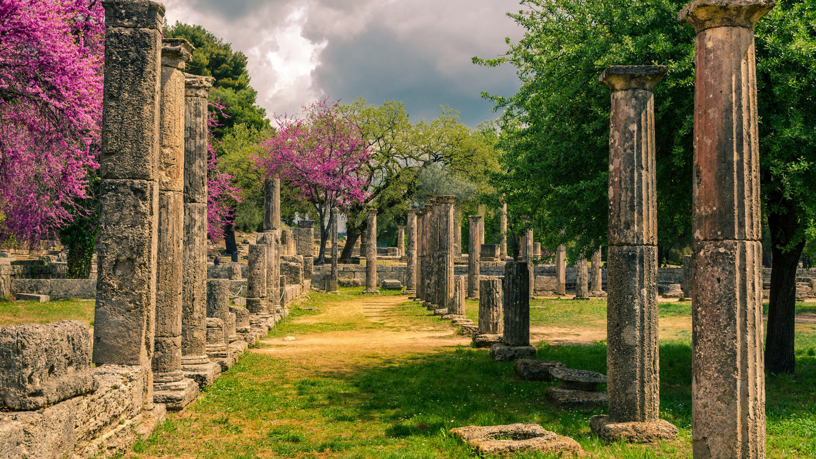 Ancient ruins of Olympia surrounded by blooming trees and greenery under a dramatic cloudy sky in Greece.