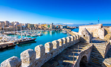 Panoramic view of Heraklion’s old Venetian harbor and fortress with boats and snow-capped mountains in the background.