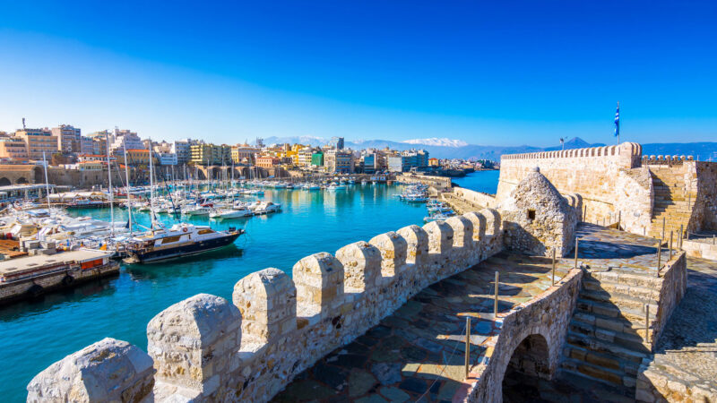 Panoramic view of Heraklion’s old Venetian harbor and fortress with boats and snow-capped mountains in the background.