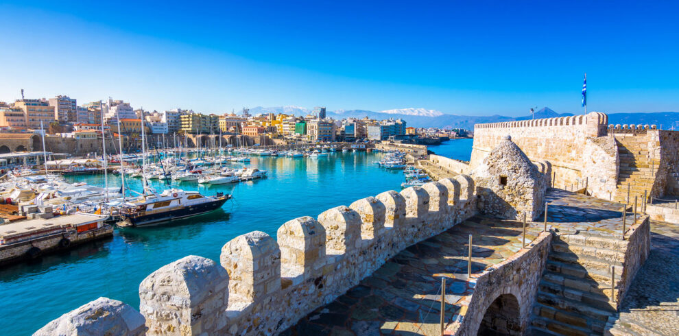 Panoramic view of Heraklion’s old Venetian harbor and fortress with boats and snow-capped mountains in the background.