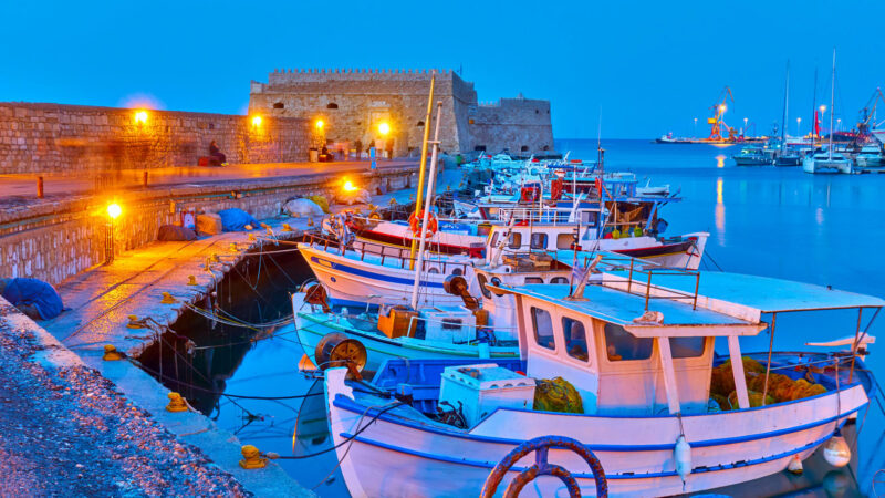 Traditional fishing boats docked in the illuminated harbor of Heraklion, Crete, with the Venetian fortress at dusk.