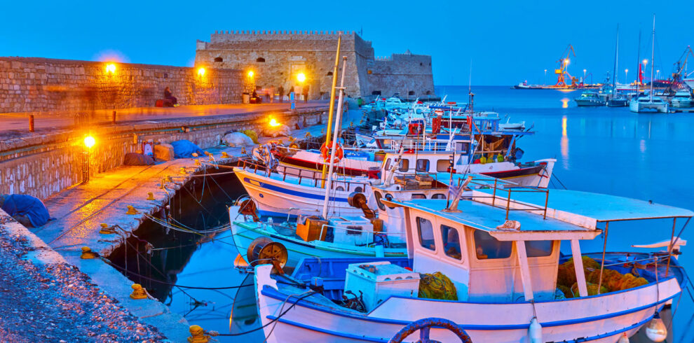 Traditional fishing boats docked in the illuminated harbor of Heraklion, Crete, with the Venetian fortress at dusk.