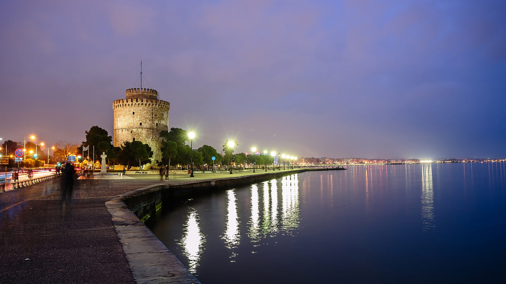 Twilight view of Thessaloniki’s White Tower and waterfront promenade reflected in the calm waters of the Thermaic Gulf.