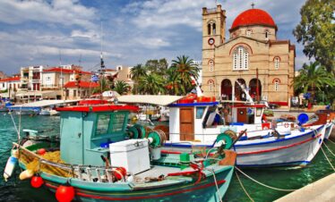 Colorful fishing boats docked in the harbor of Aegina with a red-domed church and palm trees in the background.