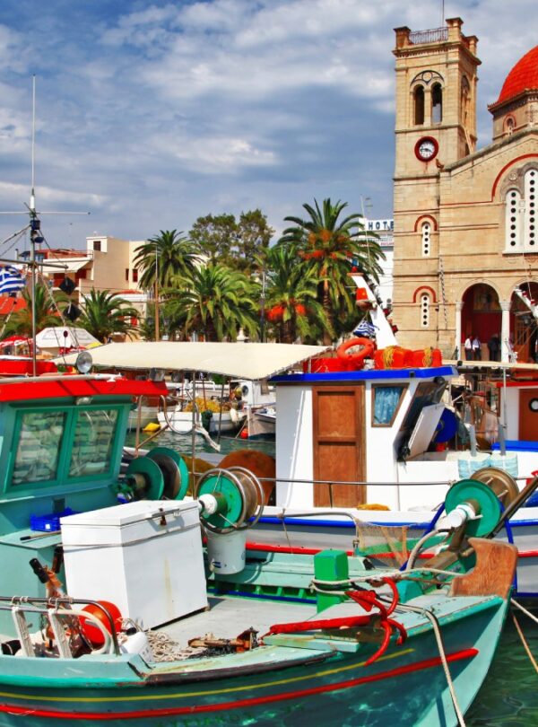 Colorful fishing boats docked in the harbor of Aegina with a red-domed church and palm trees in the background.
