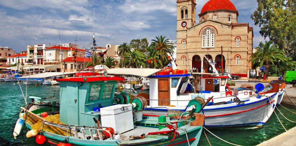 Colorful fishing boats docked in the harbor of Aegina with a red-domed church and palm trees in the background.