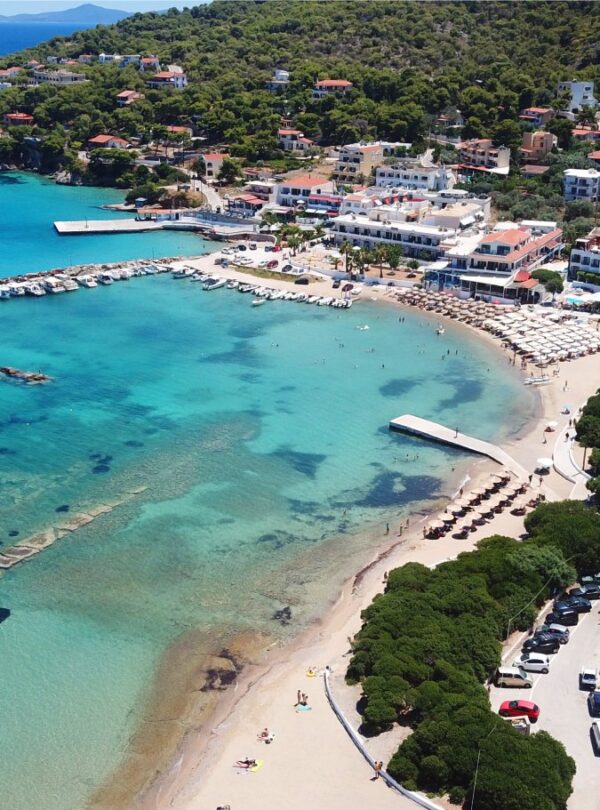 Aerial view of a picturesque Greek seaside village with turquoise waters, sandy beach, small marina, and a white church with a blue dome.