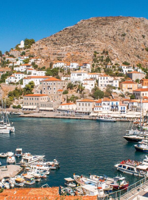 Scenic view of Hydra island harbor in Greece, with sailboats and fishing boats docked along the waterfront, traditional whitewashed houses with red-tiled roofs climbing the hillside, and rocky mountains in the background under a clear blue sky.