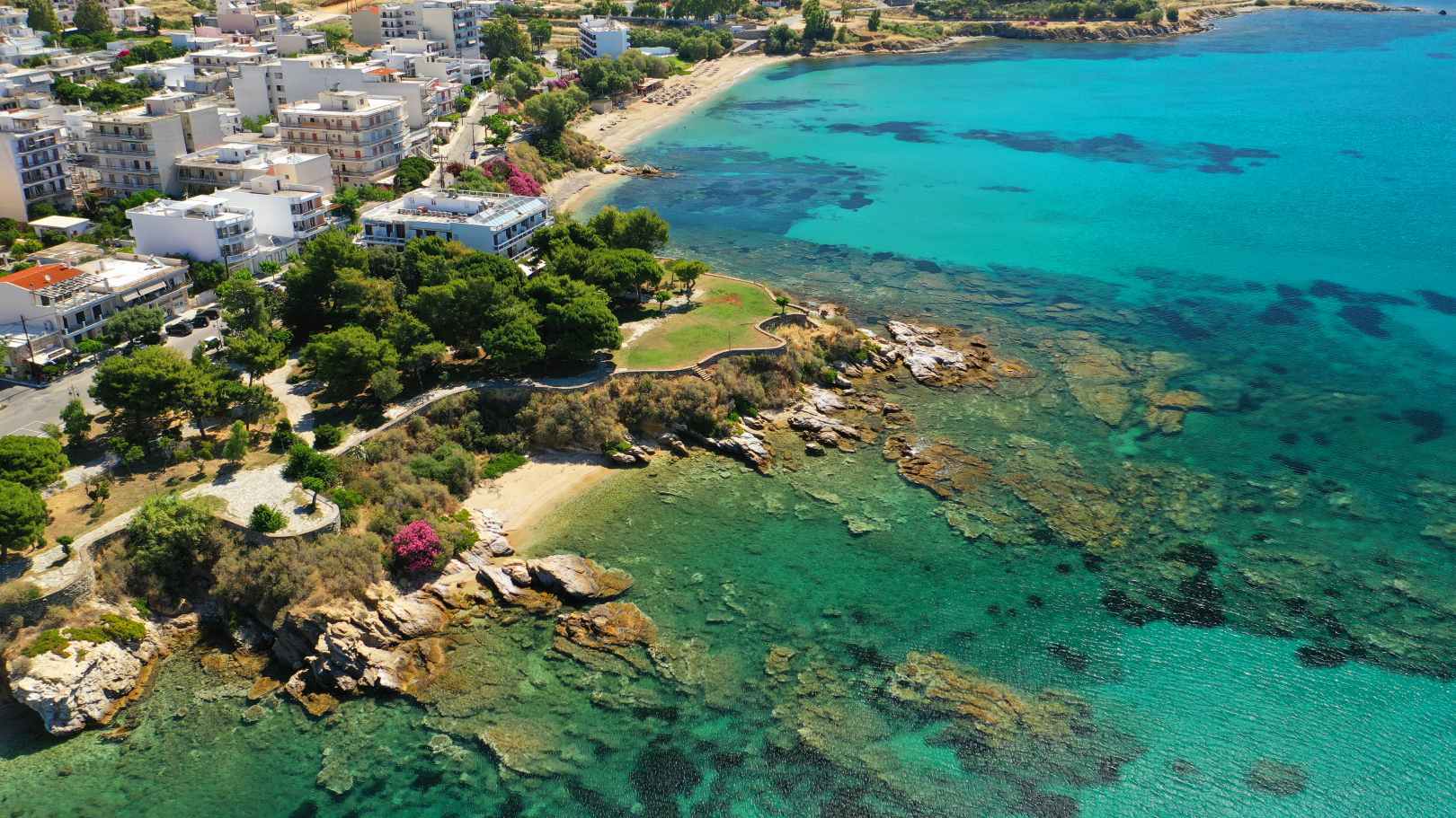 Aerial view of a scenic coastal town with turquoise waters, rocky shoreline, sandy beaches, and lush greenery blending into the urban landscape.