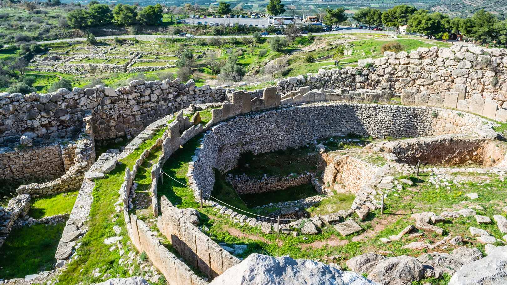 Archaeological remains of the royal tombs at Mycenae in Greece, showcasing circular grave enclosures and ancient stone walls surrounded by a scenic landscape.