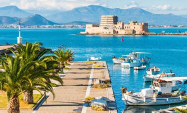 Scenic view of Nafplio’s harbor with fishing boats, palm-lined promenade, and the historic Bourtzi fortress in the sea.