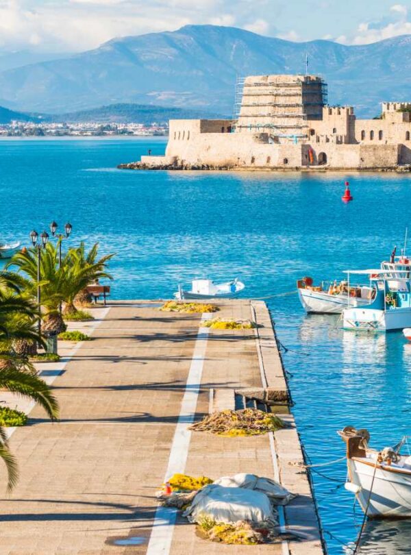 Scenic view of Nafplio’s harbor with fishing boats, palm-lined promenade, and the historic Bourtzi fortress in the sea.