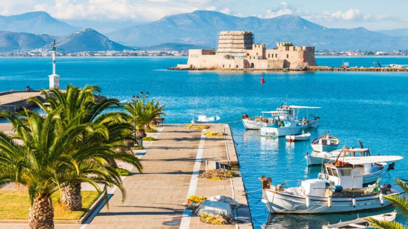 Scenic view of Nafplio’s harbor with fishing boats, palm-lined promenade, and the historic Bourtzi fortress in the sea.