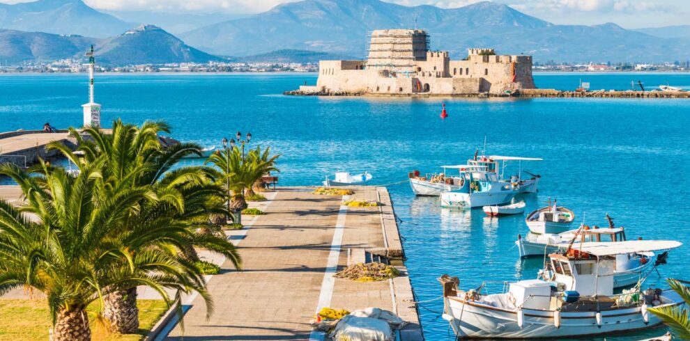 Scenic view of Nafplio’s harbor with fishing boats, palm-lined promenade, and the historic Bourtzi fortress in the sea.