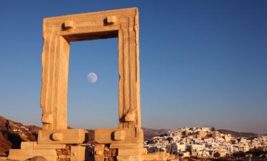 The ancient Portara gate of the Temple of Apollo on Naxos, framed against a clear blue sky with the moon rising, and the whitewashed houses of Naxos town in the background.