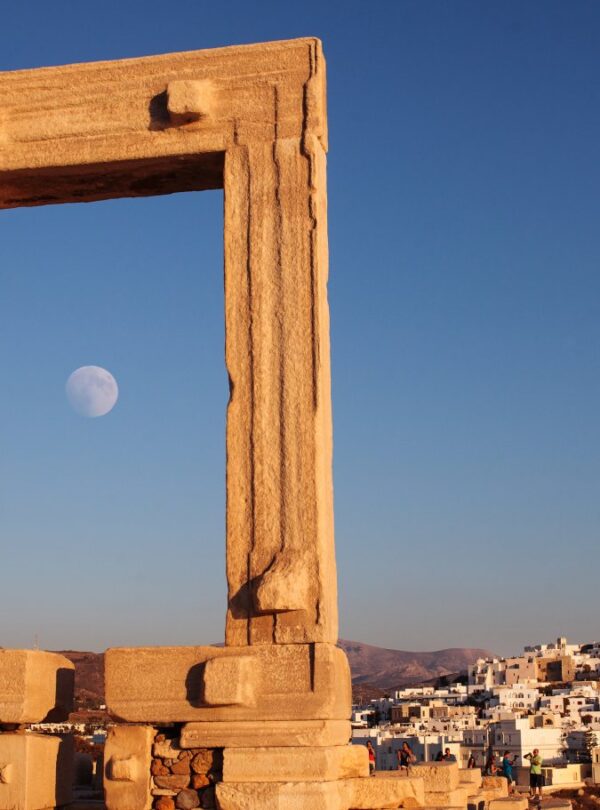 The ancient Portara gate of the Temple of Apollo on Naxos, framed against a clear blue sky with the moon rising, and the whitewashed houses of Naxos town in the background.