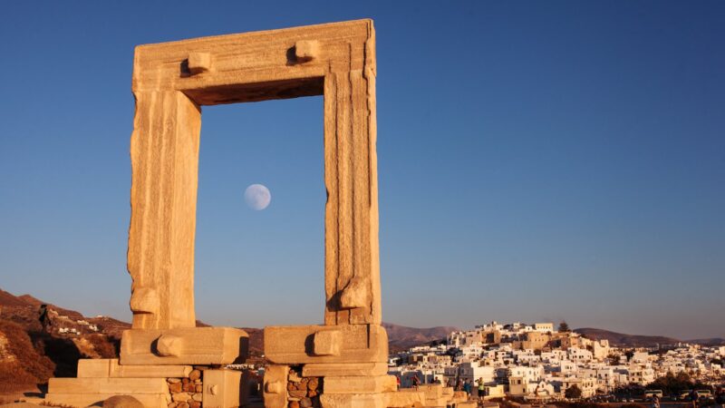 The ancient Portara gate of the Temple of Apollo on Naxos, framed against a clear blue sky with the moon rising, and the whitewashed houses of Naxos town in the background.