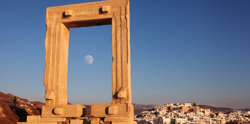 The ancient Portara gate of the Temple of Apollo on Naxos, framed against a clear blue sky with the moon rising, and the whitewashed houses of Naxos town in the background.