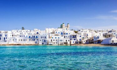 Whitewashed Cycladic houses with blue doors and windows overlooking the turquoise sea in Naoussa village on Paros island, under a clear blue sky.