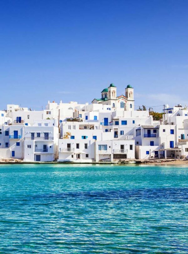 Whitewashed Cycladic houses with blue doors and windows overlooking the turquoise sea in Naoussa village on Paros island, under a clear blue sky.
