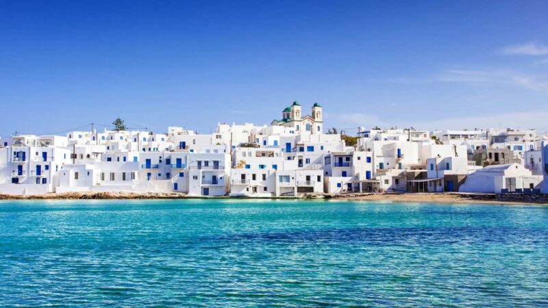 Whitewashed Cycladic houses with blue doors and windows overlooking the turquoise sea in Naoussa village on Paros island, under a clear blue sky.