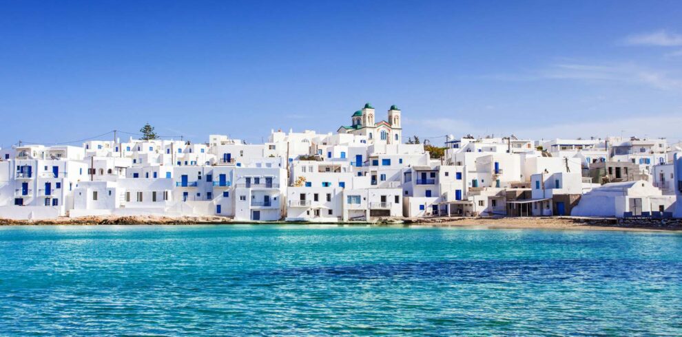 Whitewashed Cycladic houses with blue doors and windows overlooking the turquoise sea in Naoussa village on Paros island, under a clear blue sky.