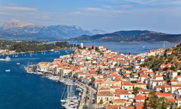 Panoramic view of Poros island, Greece, with red-roofed houses, a scenic waterfront lined with sailboats, and mountains in the distance under a clear blue sky.