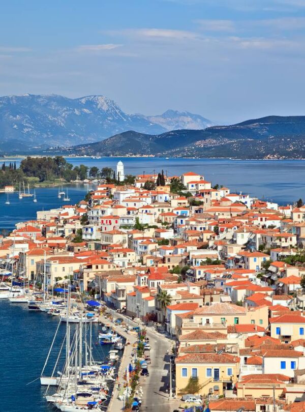 Panoramic view of Poros island, Greece, with red-roofed houses, a scenic waterfront lined with sailboats, and mountains in the distance under a clear blue sky.