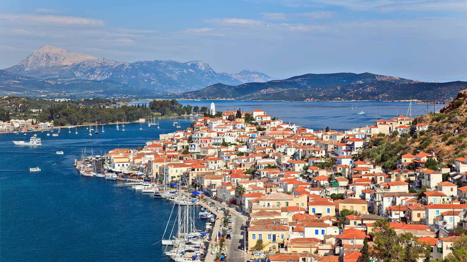 Panoramic view of Poros island, Greece, with red-roofed houses, a scenic waterfront lined with sailboats, and mountains in the distance under a clear blue sky.