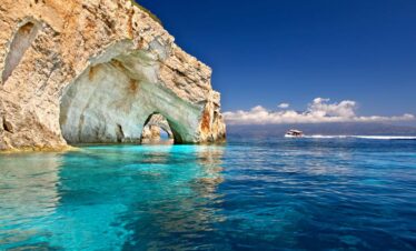 The stunning Blue Caves of Zakynthos with turquoise Ionian waters and a boat sailing under a clear blue sky.