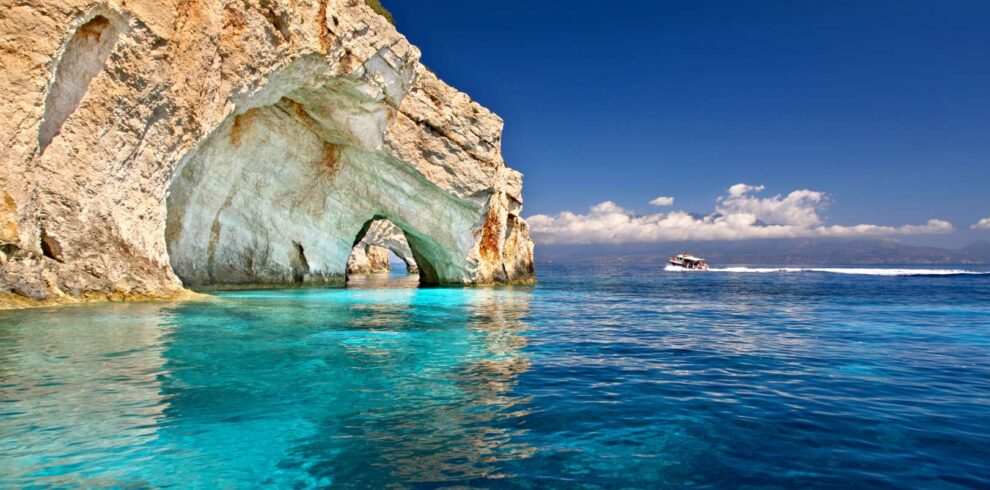 The stunning Blue Caves of Zakynthos with turquoise Ionian waters and a boat sailing under a clear blue sky.
