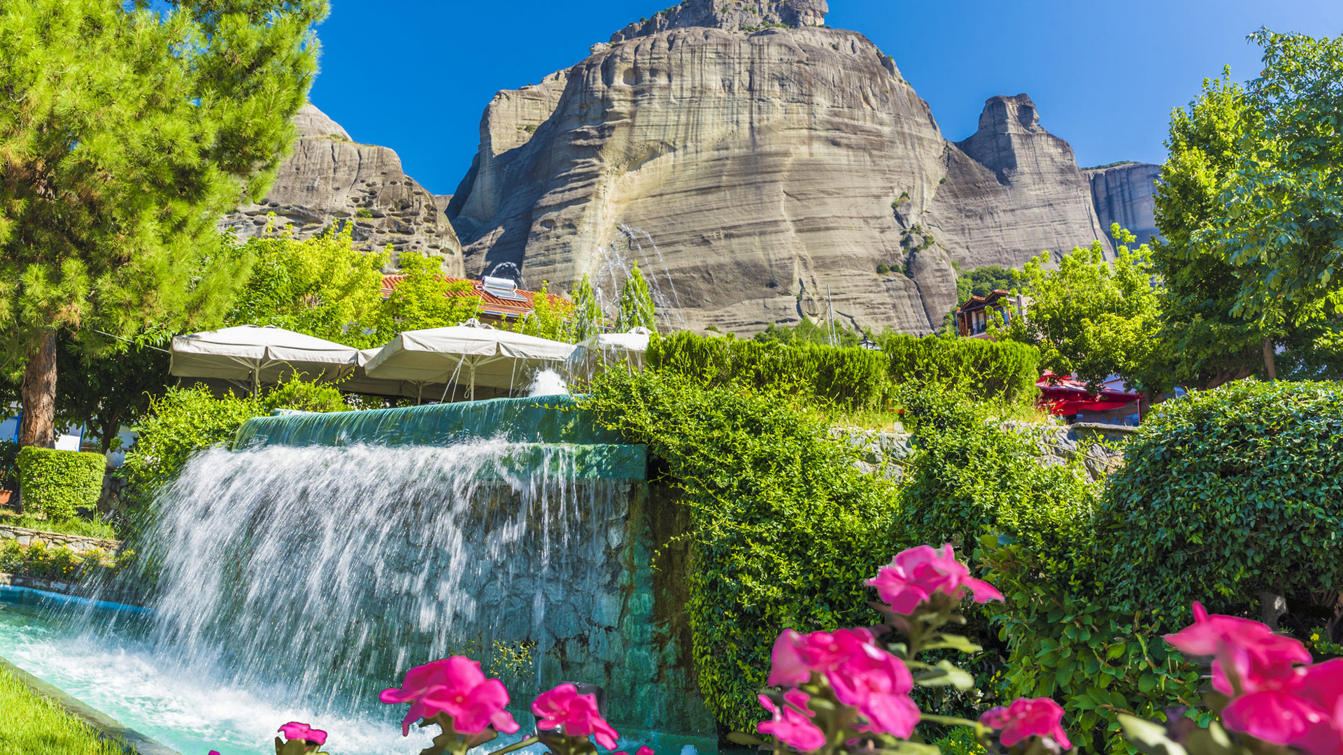 Lush garden with a waterfall and vibrant flowers set against the dramatic rock formations of Meteora, Greece.