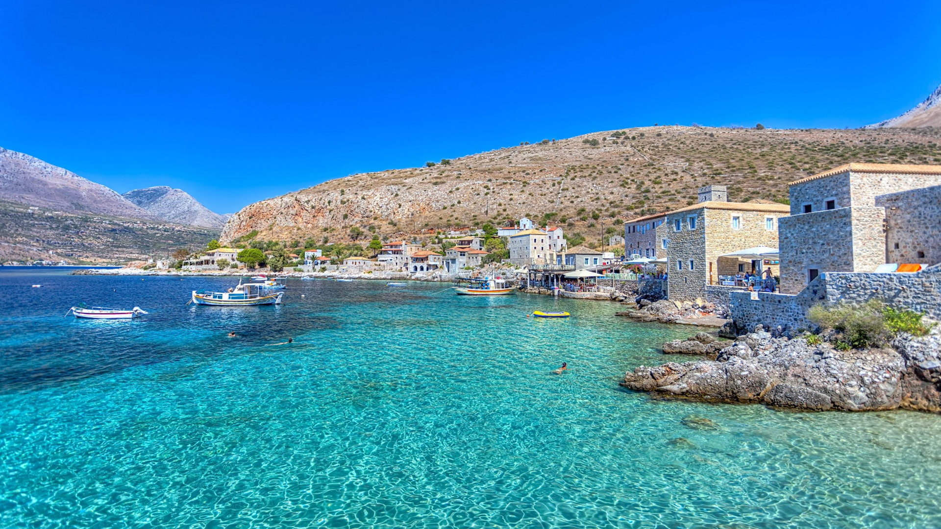 Crystal-clear turquoise waters and traditional stone houses along the coastline of Limeni in the Mani Peninsula, Peloponnese, Greece, with small fishing boats floating in the bay and rugged hills rising in the background under a bright blue sky.