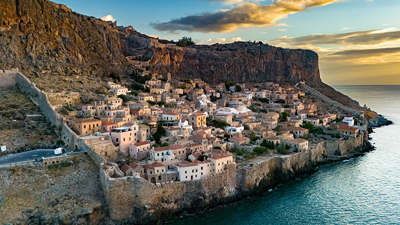 Aerial view of the medieval walled town of Monemvasia in the Peloponnese, Greece, perched on a rocky cliff overlooking the Aegean Sea, with its stone houses and narrow alleys glowing under the warm light of sunset.