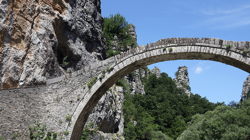 Traditional stone arch bridge in Zagori, Epirus, Greece, set against rocky cliffs and lush green landscape.