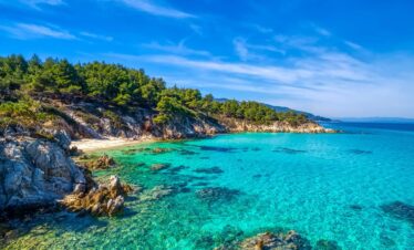Turquoise waters and pine-covered coastline of Halkidiki in northern Greece under a bright blue sky.