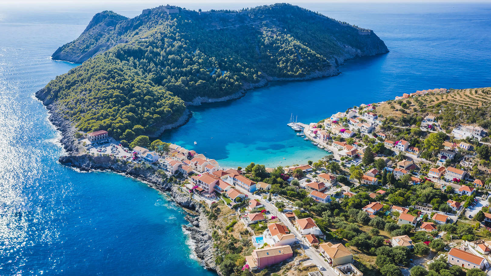 Aerial view of the picturesque Assos village on Kefalonia island, with turquoise waters, colorful houses, and lush green hills.